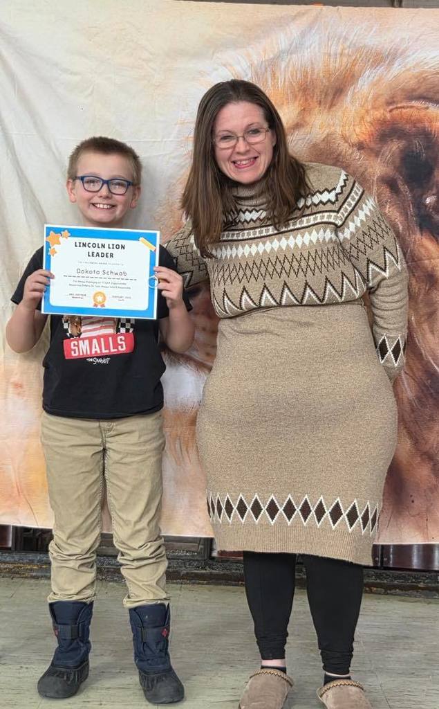 A women in a tan sweater dress smiling next to a kid  that is holding a certificate. 