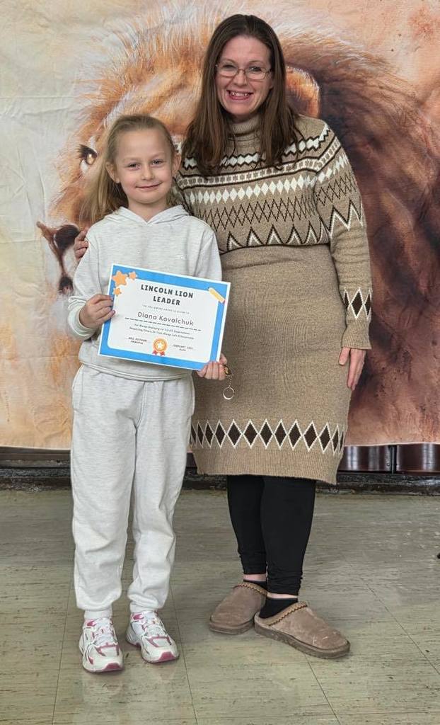 A women in a tan sweater dress smiling next to a kid  that is holding a certificate. 