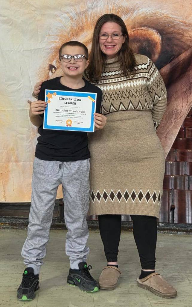 A women in a tan sweater dress smiling next to a kid  that is holding a certificate. 