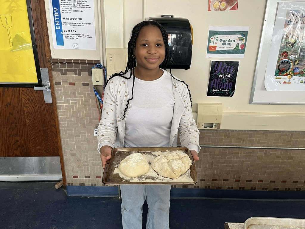A young girl in a white shirt is holding a tray with two balls of dough and one of the balls of dough has cuts in it. 