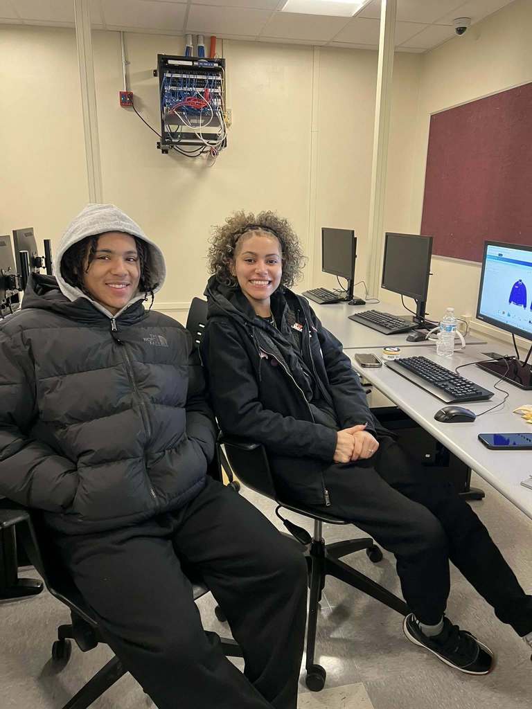 Two students wearing black jackets sitting and smiling in a computer lab.