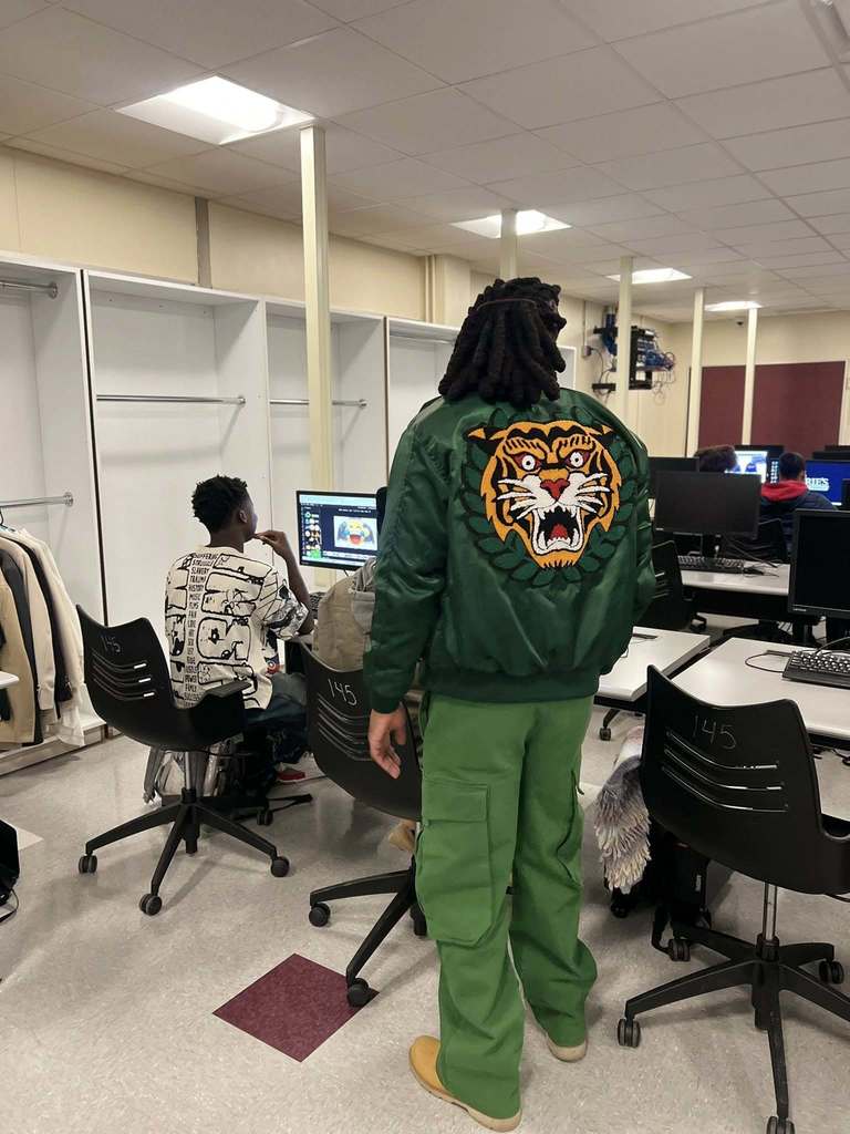 A man dressed in all green standing while students work on the computers.