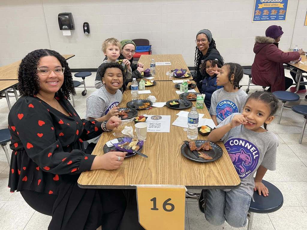 A group of adults and kids smiling and eating in a lunch cafeteria. 