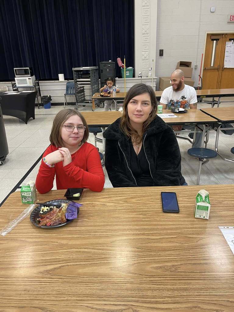A women in a black jacket and young girl in a red shirt smiling in a school cafeteria. 