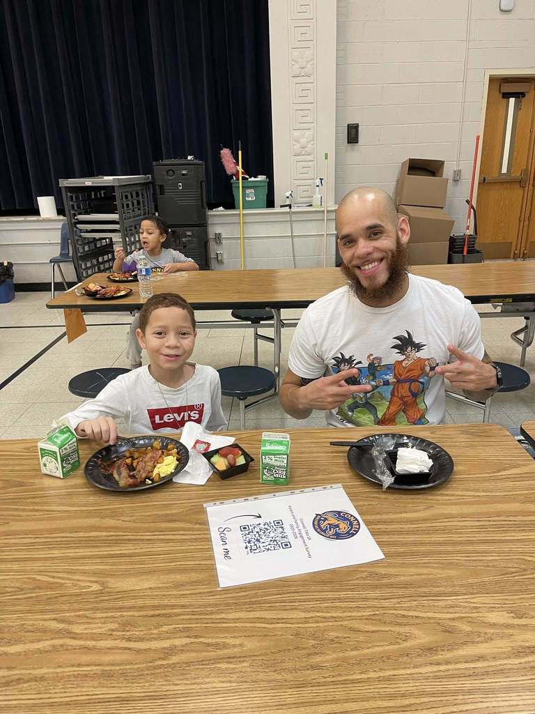 A man and young boy in white shirts smiling in a school cafeteria. 