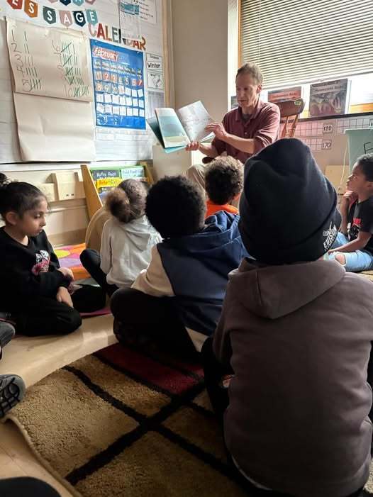 A women reading a book to a classroom of kids who are sitting on the floor. 