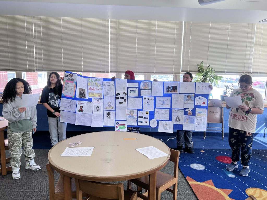 Kids holding up a long poster and reading off of papers in a classroom. 
