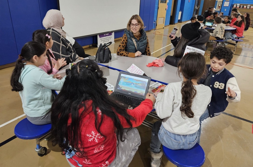 Kids and adults sitting at a circle table with laptops and colorful charts in front of them.