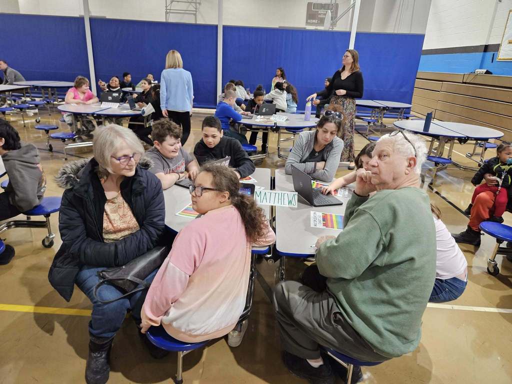 Kids and adults sitting at a circle table with laptops and colorful charts in front of them.
