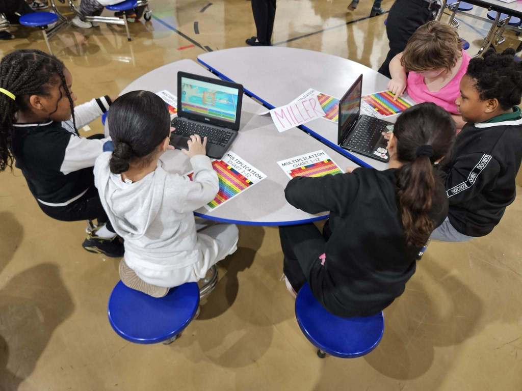 Kids sitting at a circle table with laptops and colorful charts in front of them.