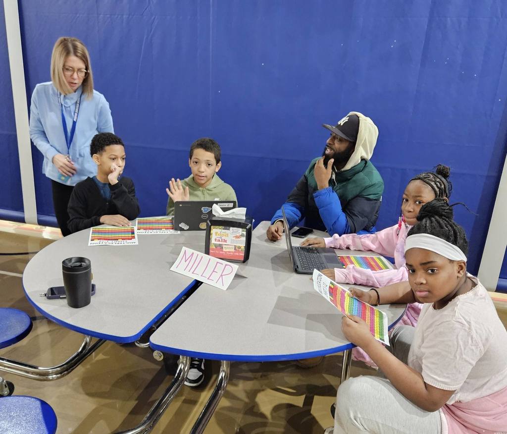 Kids and adults sitting at a circle table with laptops and colorful charts in front of them.