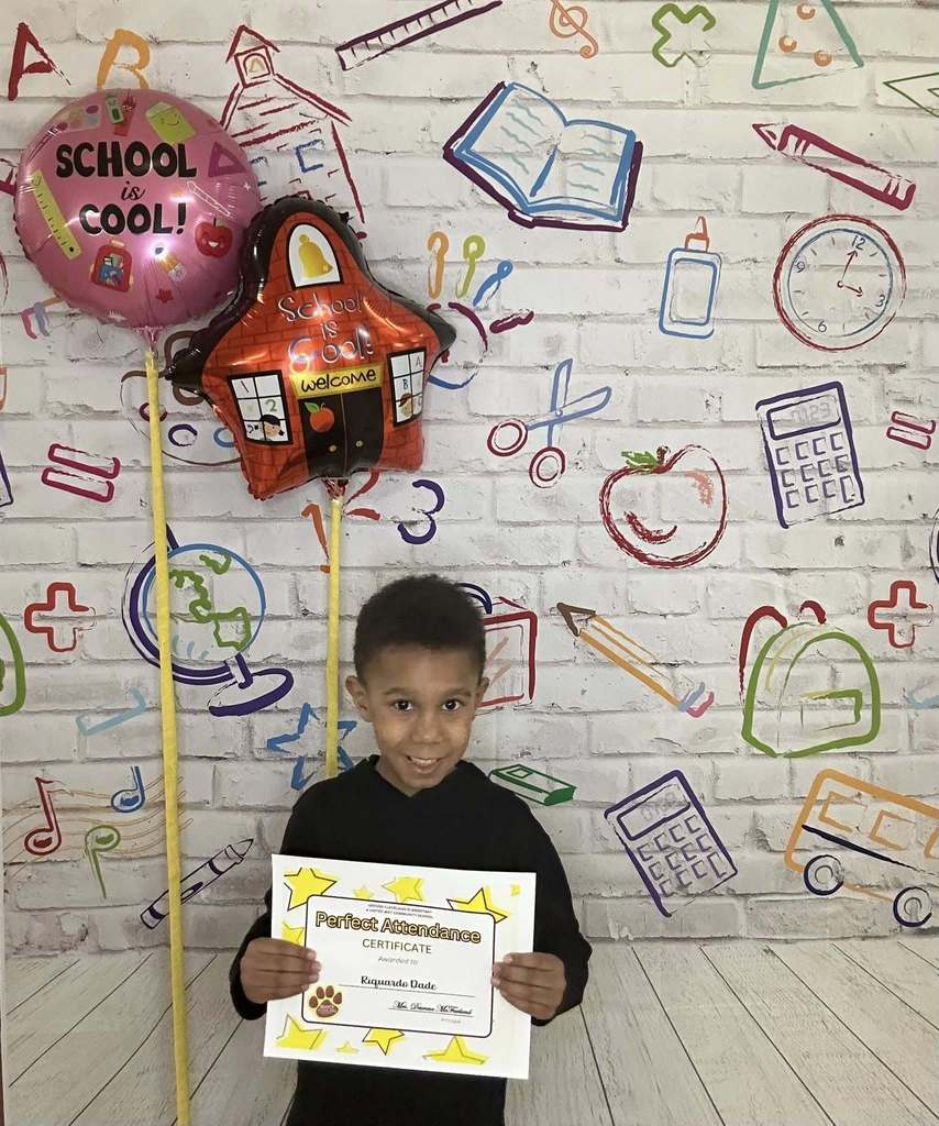 Young boy wearing a black shirt is holding his certificate in front of a white backdrop with school graphics on it.