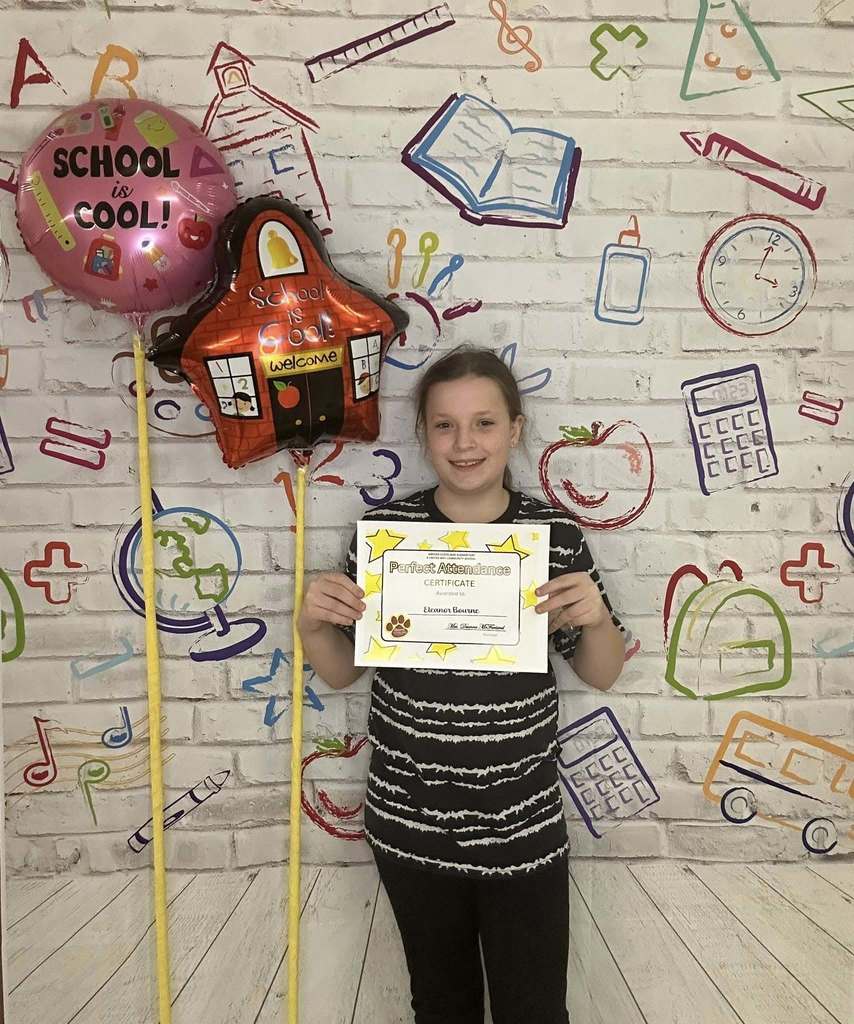Young girl wearing a black and white striped shirt is holding his certificate in front of a white backdrop with school graphics on it.