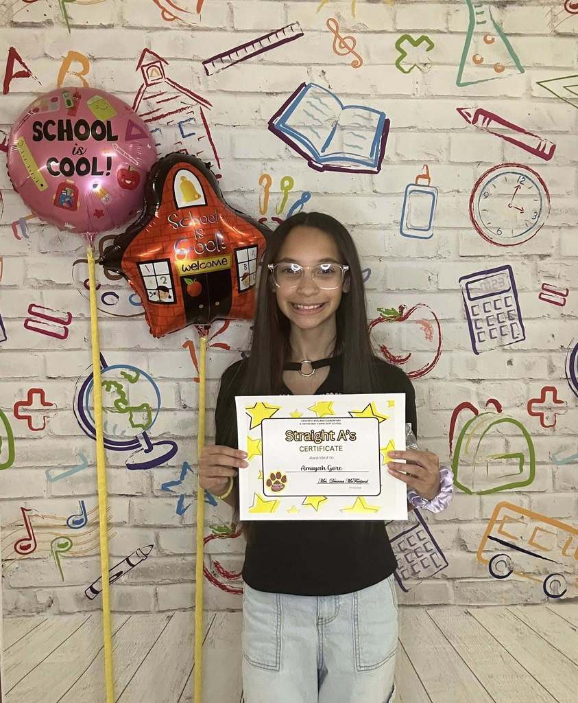 Young girl wearing a black shirt is holding his certificate in front of a white backdrop with school graphics on it.
