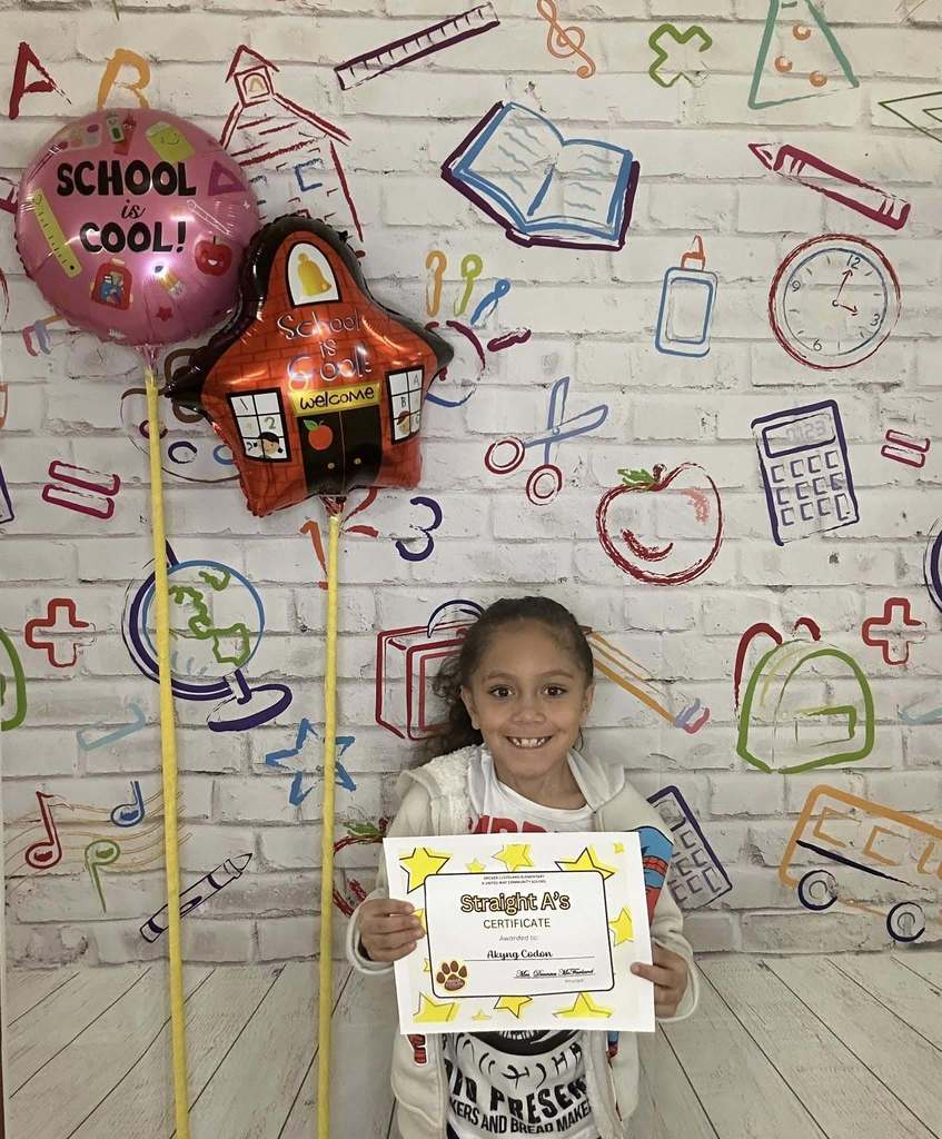 Young girl wearing a white hoodie is holding his certificate in front of a white backdrop with school graphics on it.