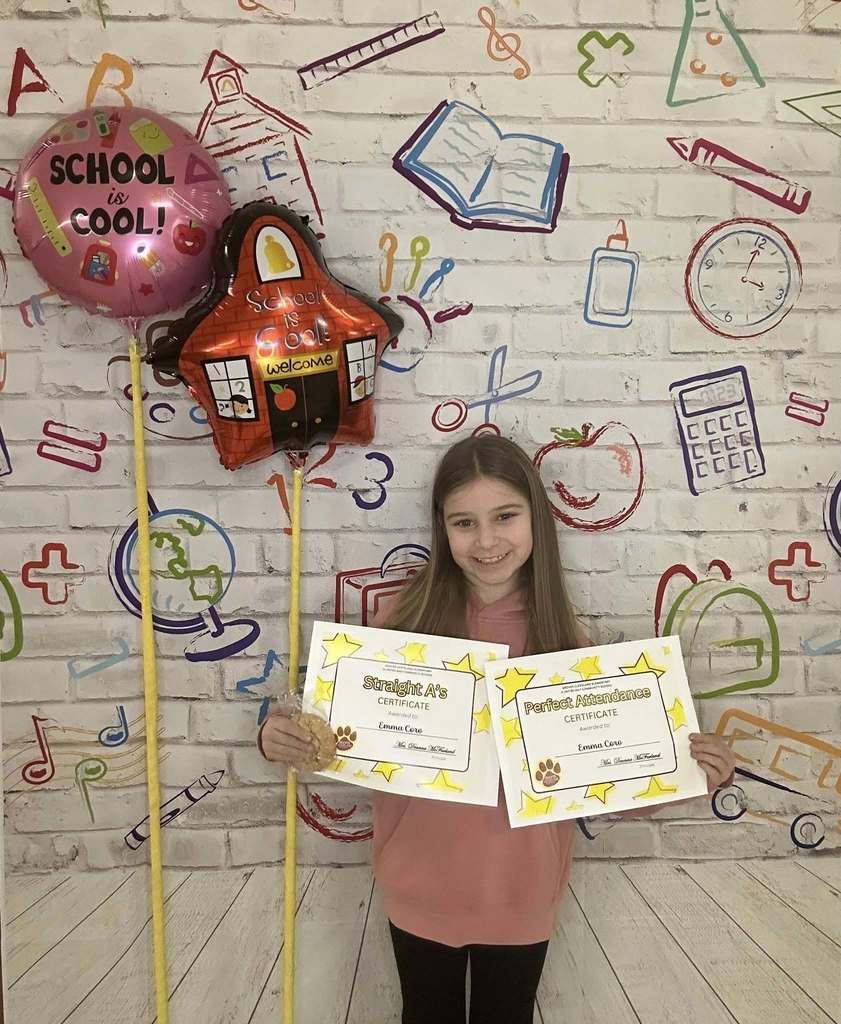 Young girl wearing a pink hoodie is holding his certificate in front of a white backdrop with school graphics on it.