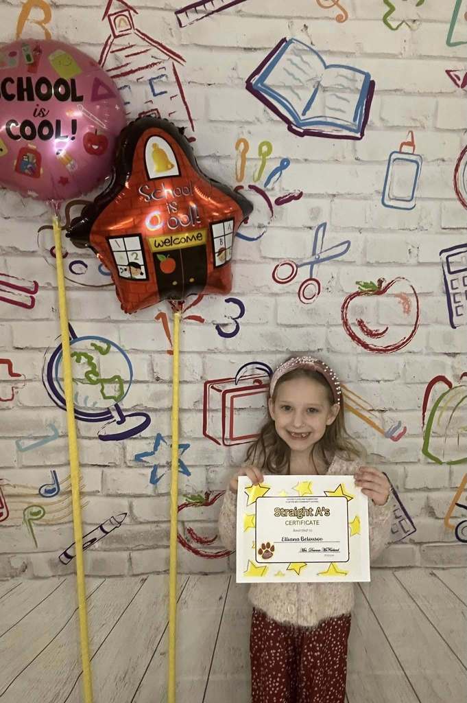 Young girl wearing red pants is holding his certificate in front of a white backdrop with school graphics on it.