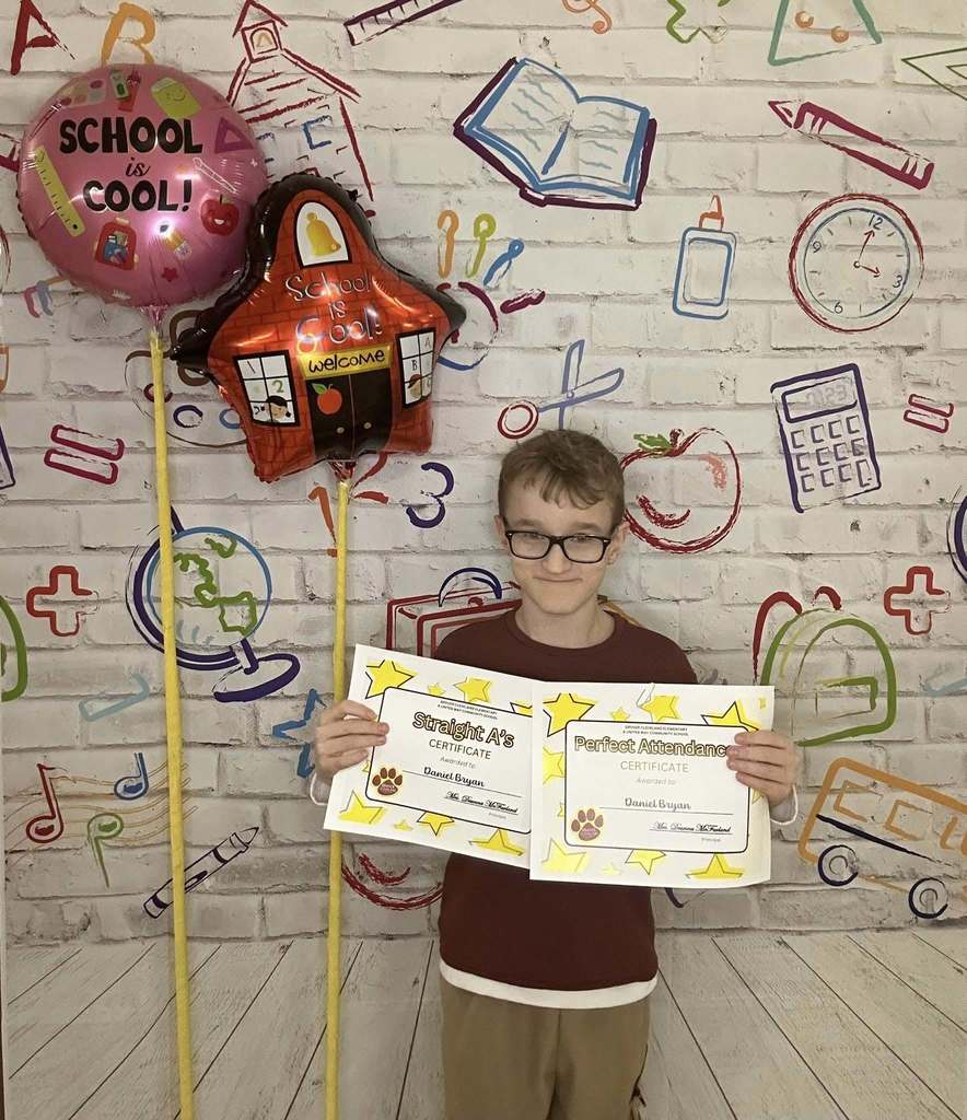 Young boy wearing a red shirt is holding his certificate in front of a white backdrop with school graphics on it.