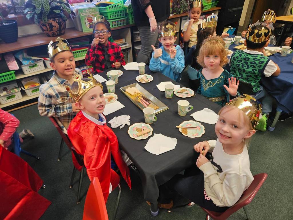 A group of kids wearing gold crowns are sitting at a table with food and drinks on the table. 