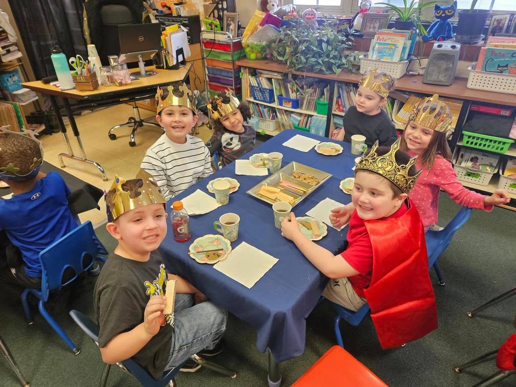 A group of kids wearing gold crowns are sitting at a table with food and drinks on the table. 