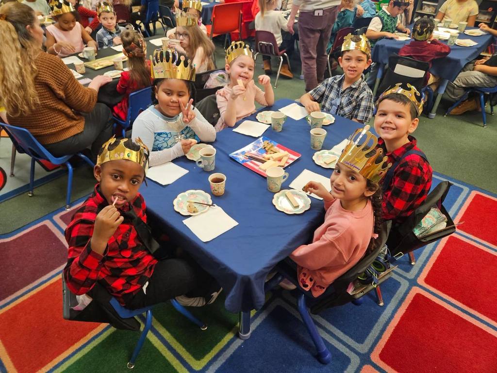 A group of kids wearing gold crowns are sitting at a table with food and drinks on the table. 