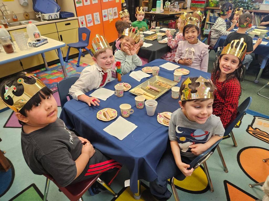 A group of kids wearing gold crowns are sitting at a table with food and drinks on the table. 