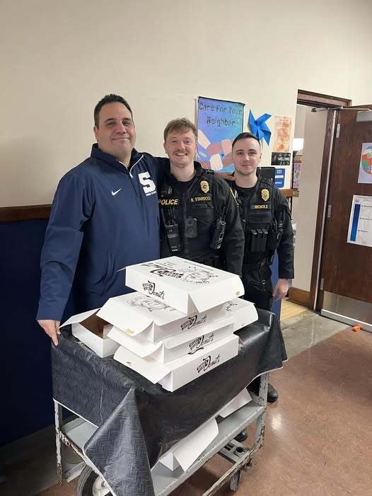 An adult with two police officers standing behind a cart that has boxes of donuts on it.