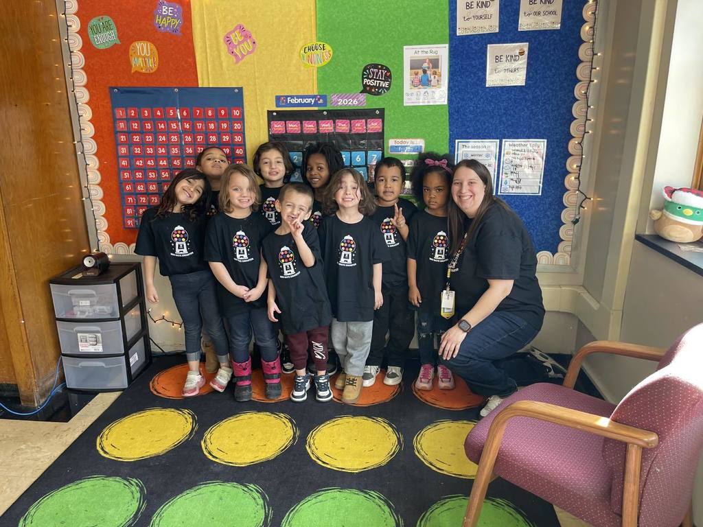 Group of kids wearing matching black shirts standing in a classroom and smiling next to their teacher. 