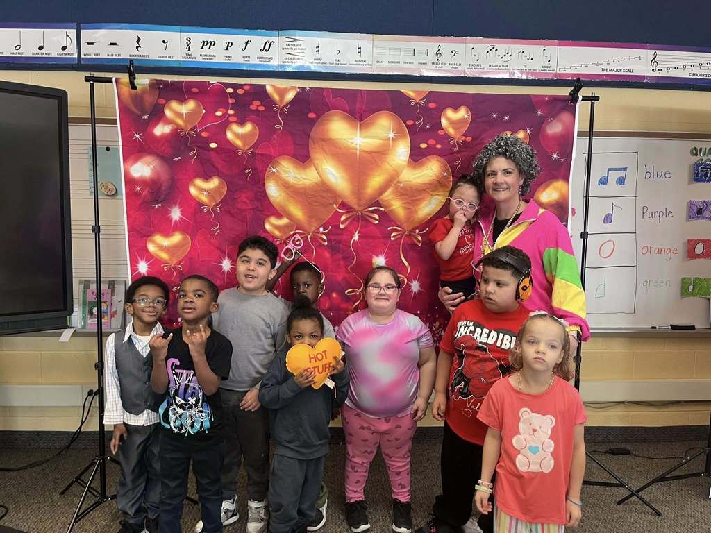 Students standing with their teacher in front of a backdrop with hearts on it.