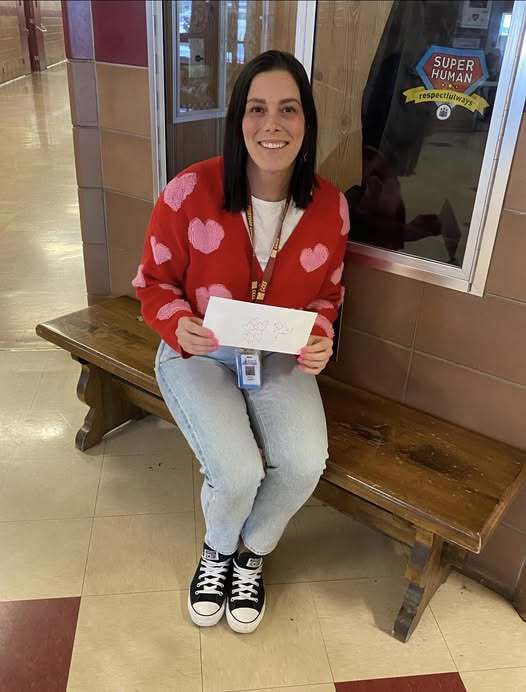 A women in a red sweater with pink hearts is sitting on a bench and holding a envelope.