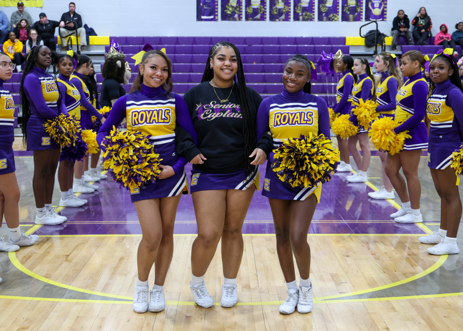 Two chearleaders standing next to a cheerleader who is wearing a senior chearleader hoodie.
