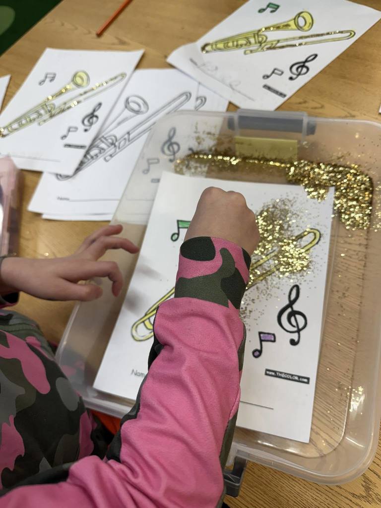 Kids putting glitter on a paper with a colored trombone on it.