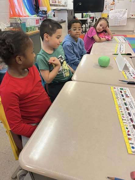 Kids sitting in class with a green ball of playdough on one table. 