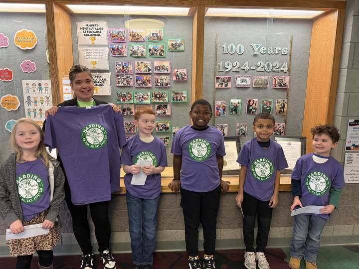 Students wearing a purple shirt with the Harding School logo on it and the teacher is holding a matching shirt. 