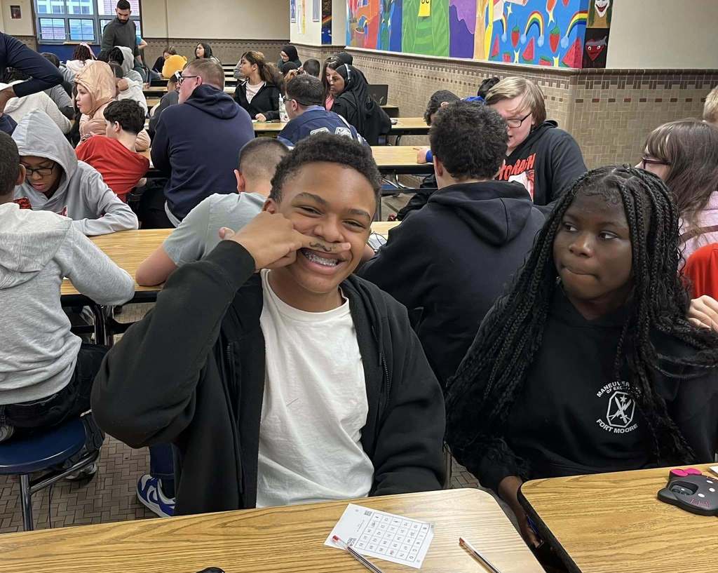 Students smiling in the school cafeteria and one boy is holding a drawn mustache on his finger to his face and smiling.