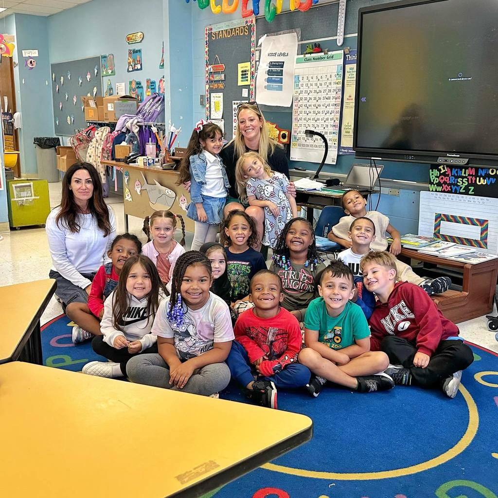 Students smiling in a classroom and there are to adults sitting with them. 