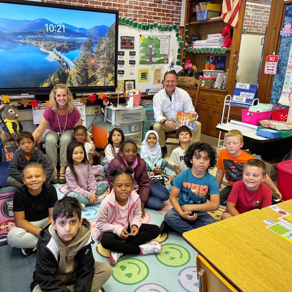 Students smiling in a classroom and there are to adults sitting with them. 