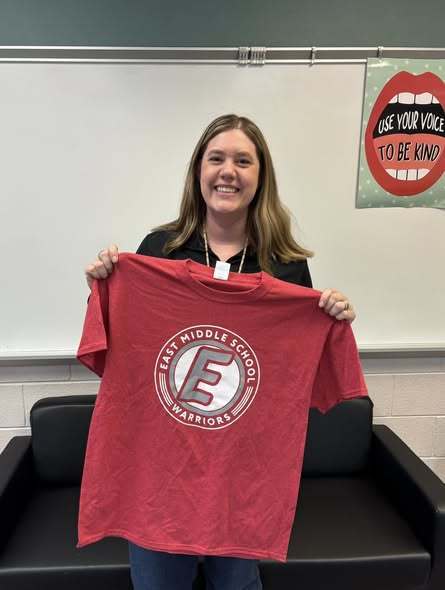A teacher holding a red shirt with the East Middle School logo on it. 