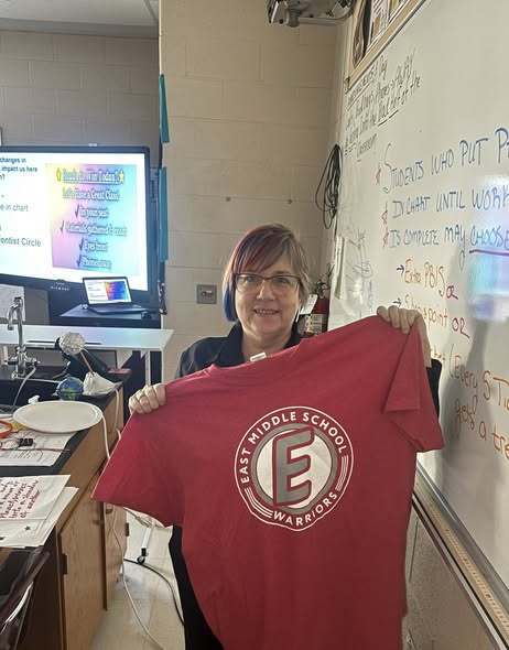 A teacher holding a red shirt with the East Middle School logo on it. 