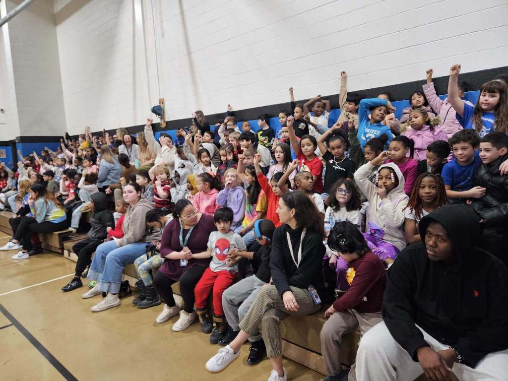 Kids raising their hands and are sitting io bleachers.