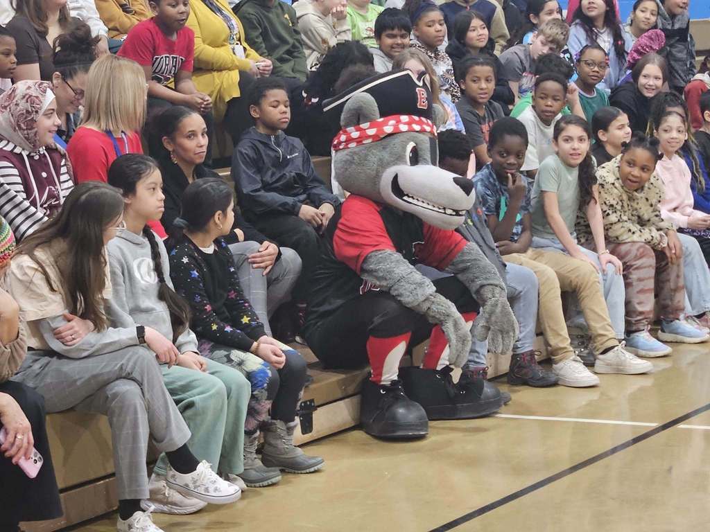 Erie SeaWolves mascot sitting next to a group of kids on the bleachers in the school gym. 
