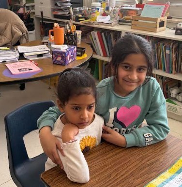 Two girls sit at a table in a classroom. One has a sunflower design on her shirt. Behind them is a bookshelf with books.