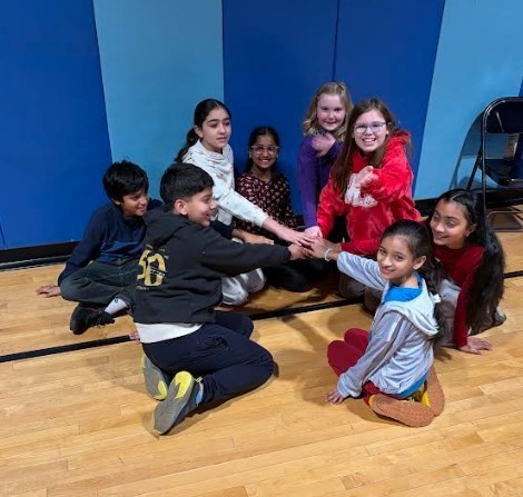 Kids in a circle holding hands, smiling, sitting on a gym floor, blue walls behind them.