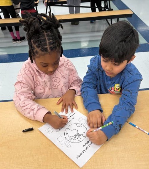 Two children in a classroom setting, one holding a pen and coloring a worksheet.