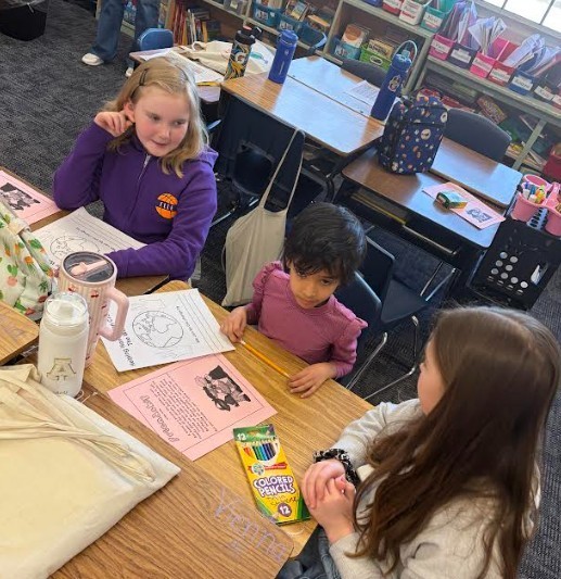 Children seated at desks with coloring books and crayons, one child holds a ruler. Background shows a classroom with shelves of books.
