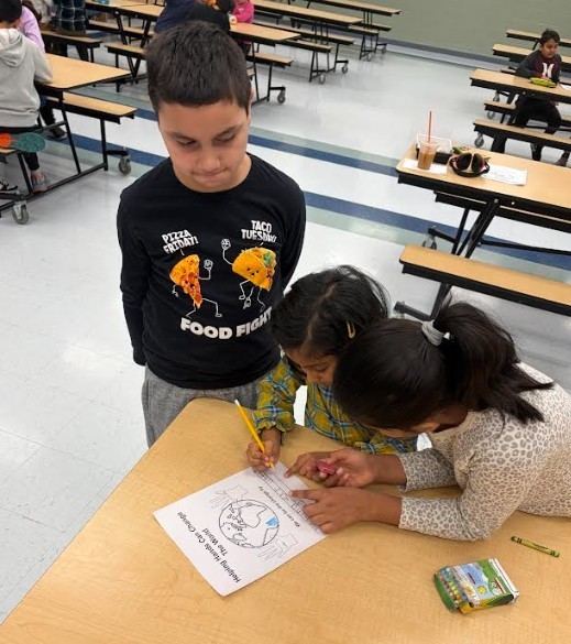 Three kids in a classroom. A boy wears a black shirt with a food design. Two girls work on a paper with a world map.