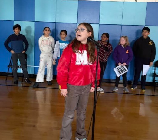 Girl in red M shirt and glasses sings into a microphone in a gym, surrounded by peers.