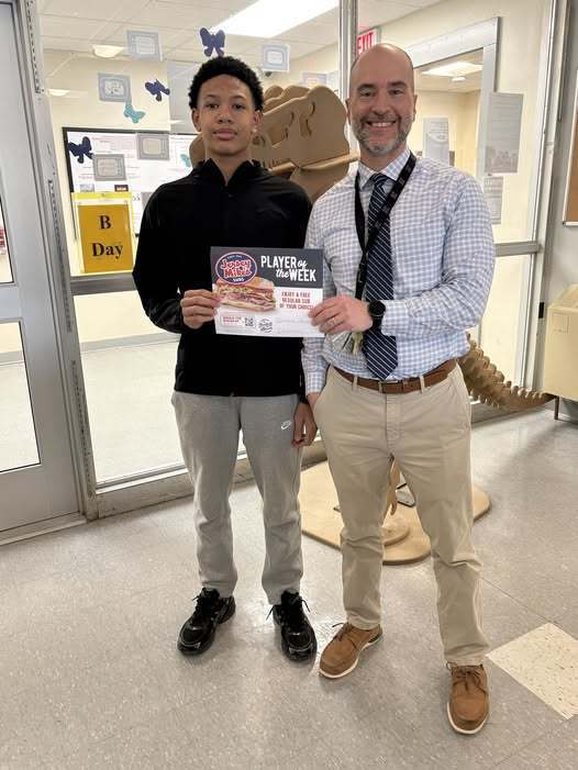 A young man in a black crewneck holding a award for player of the week with his principal who is wearing a striped tie.