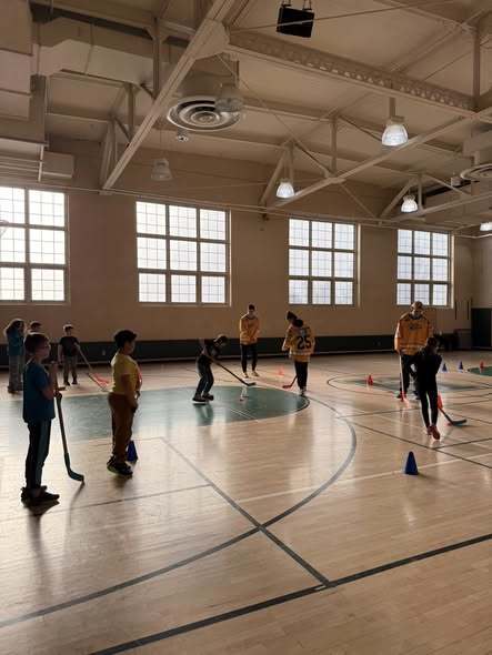 Kids playing hocky in a school gym with Erie Otters Hocky Players. 