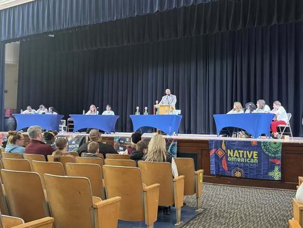 Multicultural Quiz Bowl contestants on stage at their tables while audience watches in auditorium. 
